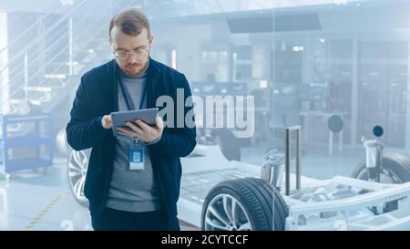 Un ingénieur de sexe masculin travaille dans un centre de développement de haute technologie qui détient une tablette. Il se trouve à côté d'un prototype de châssis de voiture électrique avec Banque D'Images