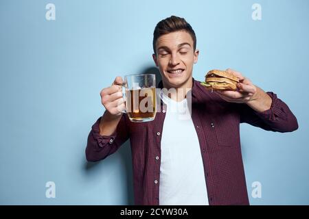 homme gai ivre avec une tasse de bière et un hamburger à la main régime alimentaire style de vie fond bleu Banque D'Images