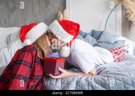 Vacances d'hiver et concept de personnes. Couple de caucasiens amoureux heureux avec cadeau de noël embrassant à la maison couché au lit portant des chapeaux de santa Banque D'Images