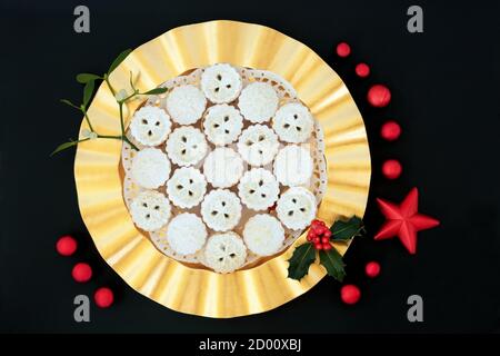 Tartes à tarte traditionnelles de Noël sur une assiette dorée avec houx aux baies d'hiver et GUI sur fond noir avec décorations de boules rouges. Banque D'Images