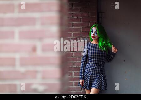 Portrait d'une fille aux cheveux verts en robe acclamée avec maquillage de joker sur un mur de brique flou d'arrière-plan. Banque D'Images