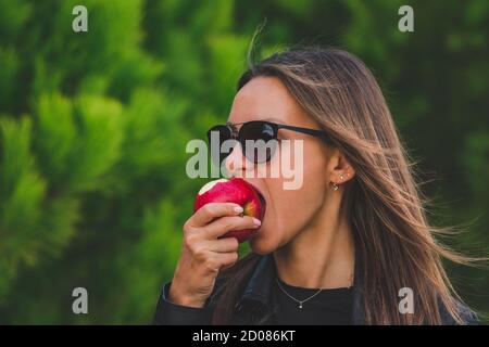 Belle jeune femme brunette vegan dans des lunettes de soleil noires mangeant la pomme rouge savoureuse sur fond d'arbres verts. Mordez la pomme. Concept de mode de vie sain. Banque D'Images