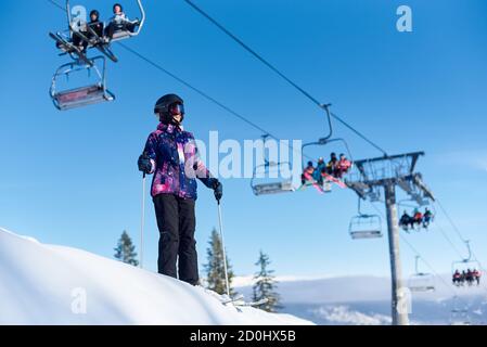 Femme souriante dans des lunettes et un casque debout sur ses skis avec des bâtons près des remontées mécaniques en action. Femme qui passe du temps à skier sur une piste de montagne enneigée. Journée ensoleillée à la station de ski. Banque D'Images