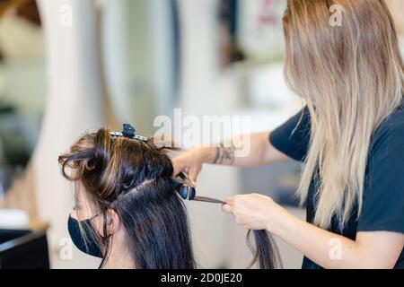 Coiffeur, protégé par un masque, peignant les cheveux de son client avec un fer à repasser dans un salon. Banque D'Images