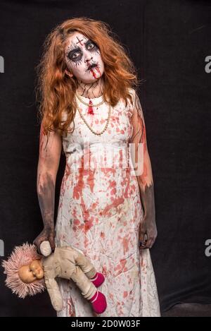 Une terrifiante tête rouge debout avec une poupée dans sa main, portant des vêtements blancs sanglants. Concept Halloween Banque D'Images