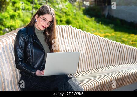 jeune femme en lunettes assise sur un banc dans le parc avec ordinateur portable et regardant la caméra lors d'une journée de printemps ensoleillée et venteuse, travaillant n'importe où aw Banque D'Images