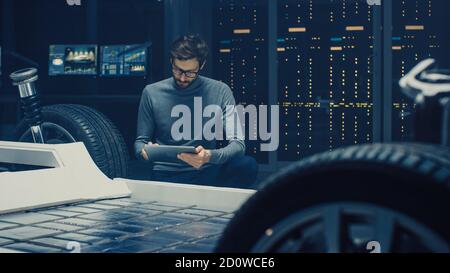 Ingénieur concepteur automobile assis à côté du prototype de plate-forme de châssis de voiture électrique hybride, utilisant un ordinateur tablette pour l'amélioration de la conception. Installation avec Banque D'Images