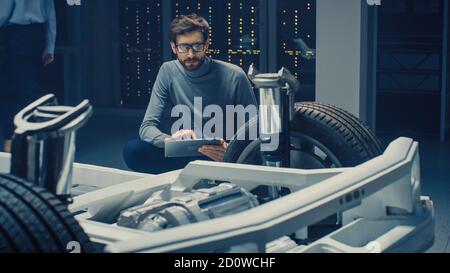Ingénieur concepteur automobile assis à côté du prototype de plate-forme de châssis de voiture électrique hybride, utilisant un ordinateur tablette pour l'amélioration de la conception. Installation avec Banque D'Images