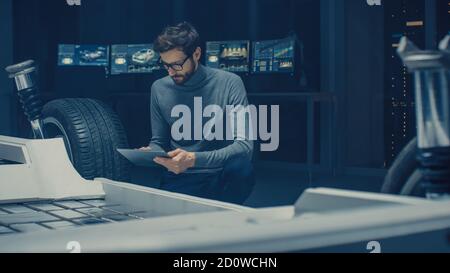 Ingénieur concepteur automobile assis à côté du prototype de plate-forme de châssis de voiture électrique hybride, utilisant un ordinateur tablette pour l'amélioration de la conception. Installation avec Banque D'Images