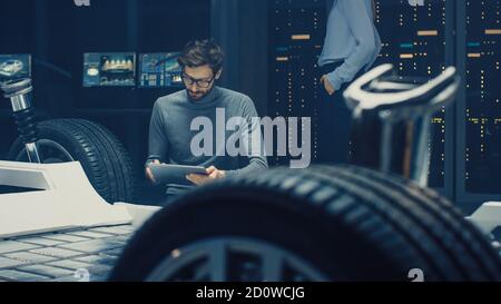 Ingénieur concepteur automobile assis à côté du prototype de plate-forme de châssis de voiture électrique hybride, utilisant un ordinateur tablette pour l'amélioration de la conception. Installation avec Banque D'Images