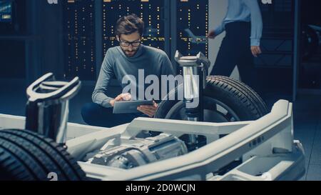 Ingénieur concepteur automobile assis à côté du prototype de plate-forme de châssis de voiture électrique hybride, utilisant un ordinateur tablette pour l'amélioration de la conception. Installation avec Banque D'Images