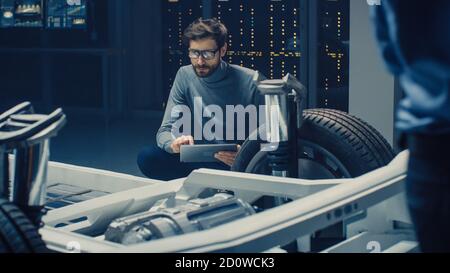 Ingénieur concepteur automobile assis à côté du prototype de plate-forme de châssis de voiture électrique hybride, utilisant un ordinateur tablette pour l'amélioration de la conception. Installation avec Banque D'Images