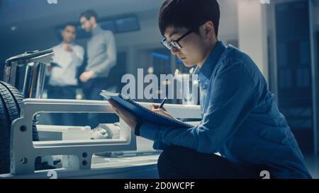 Le concepteur automobile assis à côté de la plate-forme hybride de châssis de voiture électrique, écrit des estimations et des esquisses dans le bloc-notes. Installation avec châssis de véhicule Banque D'Images