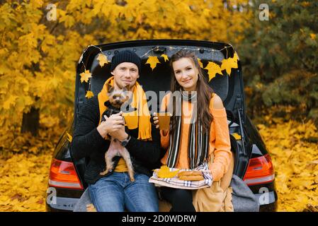 Un couple heureux et souriant de voyageurs boivent du café ou du thé tout en s'asseyant sur le coffre d'une voiture dans la forêt d'automne avec leur animal de compagnie, un chien Banque D'Images