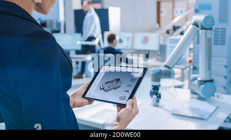 L'ingénieure féminine en robotique industrielle utilise une tablette informatique avec logiciel de CAO pour manipuler et programmer le bras robot pour soulever et déplacer des objets Banque D'Images