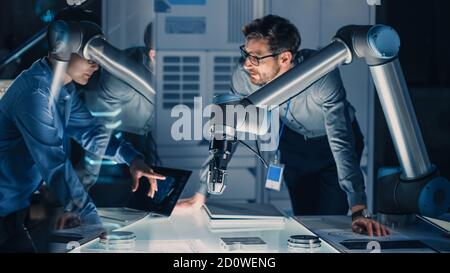 Équipe diversifiée d'ingénieurs avec ordinateur portable et tablette analyser et discuter comment un bras robotique futuriste fonctionne et déplace un objet métallique. Ils sont dans un haut Banque D'Images
