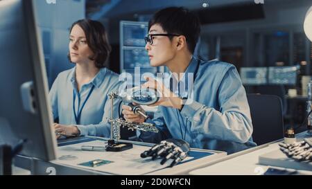 L'ingénieur japonais en développement Blue shirt sourie une carte de circuit dans un laboratoire de recherche de haute technologie avec des équipements modernes. Son collègue le demande Banque D'Images