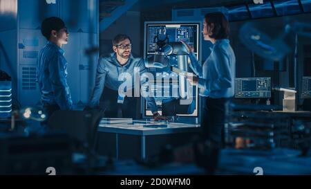 Équipe diversifiée d'ingénieurs avec ordinateur portable et tablette analyser et discuter comment un bras robotique futuriste fonctionne et déplace un objet métallique. Ils sont dans un haut Banque D'Images
