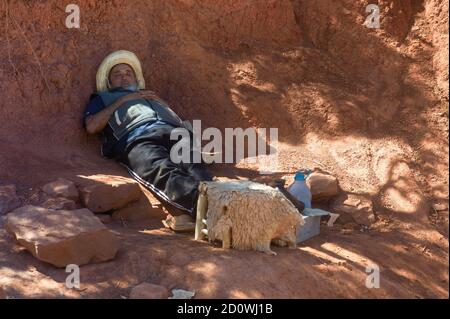 Berber homme avec chapeau de paille blanc repose dans le rouge sable à l'ombre dans le désert en attendant les clients pour une promenade à dos de chameau Banque D'Images