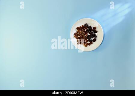 Fruits secs ensoleillés, raisins secs clairs et foncés, sur une soucoupe blanche ronde en céramique, sur fond bleu. Banque D'Images