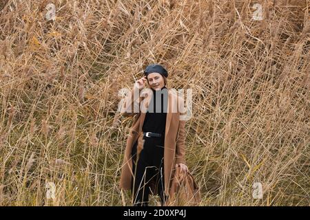 Romantique femme brune dans un béret noir brillant et un manteau de laine beige posant contre un champ dans un champ étonnant, sur la forêt de fond. Couleurs chaudes Banque D'Images