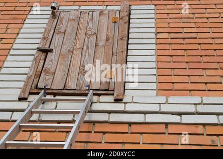 Ancienne porte en bois au grenier sur charnières rouillées, fermée avec verrou de plaque tournante et haut de l'échelle en métal adossé à elle sur fond de mur de brique et toit pignon Banque D'Images