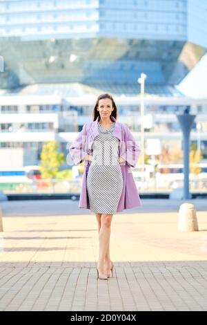 Femme tendance en manteau lilas et robe rayée posant à l'extérieur Banque D'Images