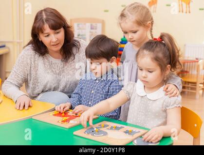 le professeur de l'école primaire recueille le puzzle avec trois élèves assis à la table. Une fille debout derrière Banque D'Images