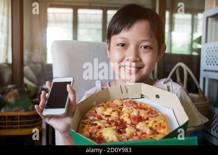 Petite fille asiatique tenant un smartphone et commandant de la pizza à manger à la maison Banque D'Images