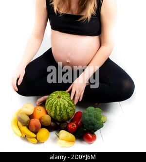 Femme enceinte avec des fruits et des légumes autour d'elle. Concept de saine alimentation. Grossesse, petite fille fine en haut noir et leggings isolés sur le blanc b. Banque D'Images