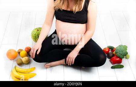 Femme enceinte avec des fruits et des légumes autour d'elle. Concept de saine alimentation. Grossesse, petite fille fine en haut noir et leggings isolés sur le blanc b. Banque D'Images