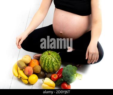 Femme enceinte avec des fruits et des légumes autour d'elle. Concept de saine alimentation. Grossesse, petite fille fine en haut noir et leggings isolés sur le blanc b. Banque D'Images