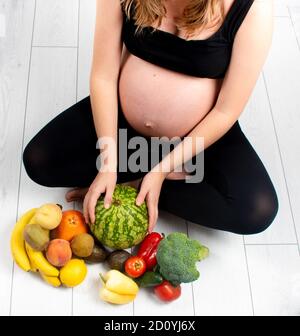 Femme enceinte avec des fruits et des légumes autour d'elle. Concept de saine alimentation. Grossesse, petite fille fine en haut noir et leggings isolés sur le blanc b. Banque D'Images