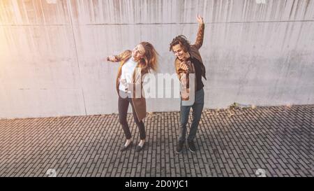 Gai Girl et Happy Young Man avec de longs cheveux dansent activement sur une rue à côté d'un mur en béton urbain. Ils portent une veste en cuir marron et Banque D'Images