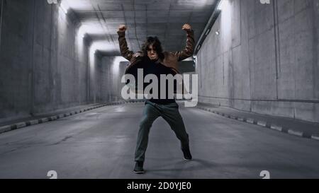 Serious Cool Young Hipster Man with long Hair est énergique Dancing Hip Hop dans un tunnel de béton éclairé. Il porte une veste en cuir marron. Banque D'Images
