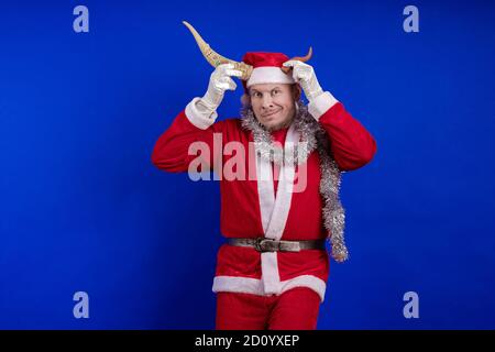 Un acteur masculin dans un costume du Père Noël tient des cornes, des blagues, des grimaces et pose sur un fond bleu Banque D'Images