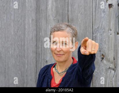 Femme de campagne caucasienne d'âge moyen déterminée avec de courts points de cheveux au spectateur avec l'index de la main gauche, en face de l'arrière-plan en bois de la grange ancienne. Banque D'Images
