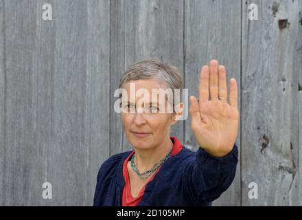 Femme de campagne caucasienne d'âge moyen sérieuse fait le panneau stop de paume de la main avec sa main gauche (geste d'arrêt), en face de vieux fond de bois de grange. Banque D'Images