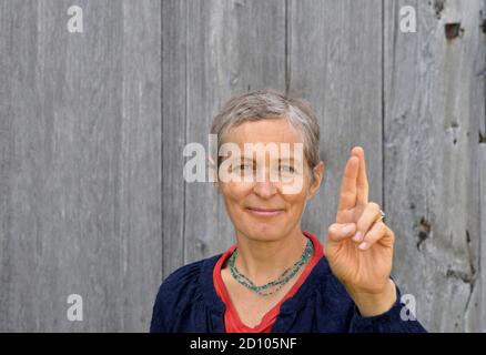 Une femme de campagne blanche d'âge moyen positive avec des cheveux courts garde ses doigts croisés et souhaite bonne chance, devant un arrière-plan en bois de la grange ancienne. Banque D'Images