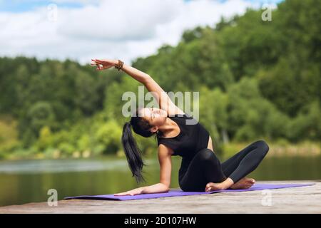 Jeune femme en vêtements de sport noirs sur un pont en bois par le lac pratiquant des asanas de yoga Banque D'Images