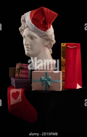 Statue en plâtre blanc d'Apollo Belvedere dans une casquette rouge du Père Noël, boîtes cadeaux et paquets sur fond coloré. Composition pour félicitations Banque D'Images