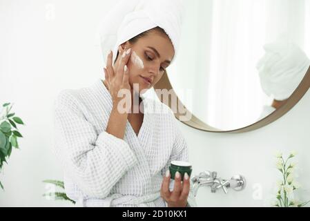 Belle jeune femme appliquant de la crème dans la salle de bains. Crème pour le visage. Soins de la peau. Belle femme souriante avec une serviette sur la tête après la douche. Banque D'Images