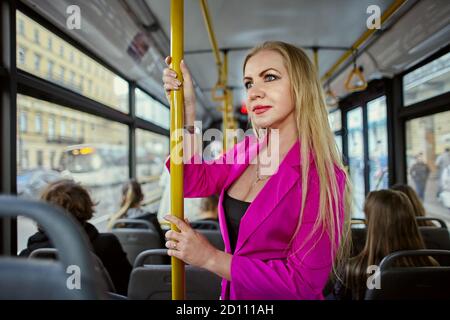 Femme caucasienne vêtue de rose, se tient à l'intérieur d'un bus ou d'un trolleybus. Des manèges blancs féminins en se tenant dans la cabine des transports en commun et h Banque D'Images