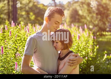 Un jeune homme souriant embrasse une fille triste en datant dans un parc public à la fin d'une journée ensoleillée d'été. Couple caucasien pour une promenade dans le jardin de la ville. Banque D'Images