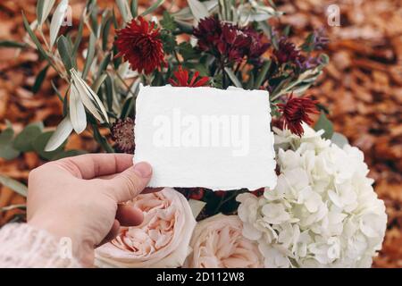 Mariage d'automne, ambiance de maquette d'anniversaire, composition avec branches d'olive et fleurs roses. Gros plan des mains de la femme tenant une carte de vœux vierge Banque D'Images