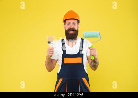 Happy Painter dans les vêtements de travail tient rouleau de peinture et brosse outils de peinture pour le bâtiment et les travaux de construction, la finition. Banque D'Images