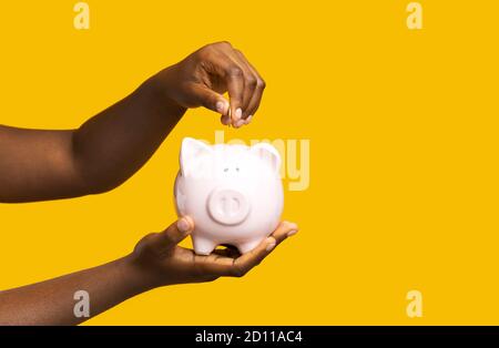 Économies d'argent. Femme noire méconnue mettant de la pièce dans la pigegybank sur fond jaune Banque D'Images