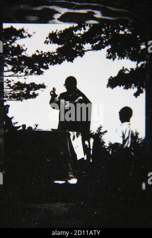 Belle photographie en noir et blanc vintage des années 1970 d'un prêtre catholique romain célébrant la Sainte Messe avec l'Eucharistie dans les champs ouverts. Banque D'Images