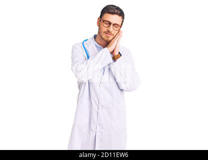 Beau jeune homme avec un ours portant un uniforme de médecin dormant fatigué rêvant et posant avec les mains ensemble tout en souriant avec les yeux fermés. Banque D'Images