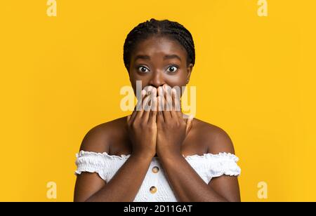 Choqué Afro-américain femelle couvrant la bouche avec les mains, posant sur fond jaune Banque D'Images